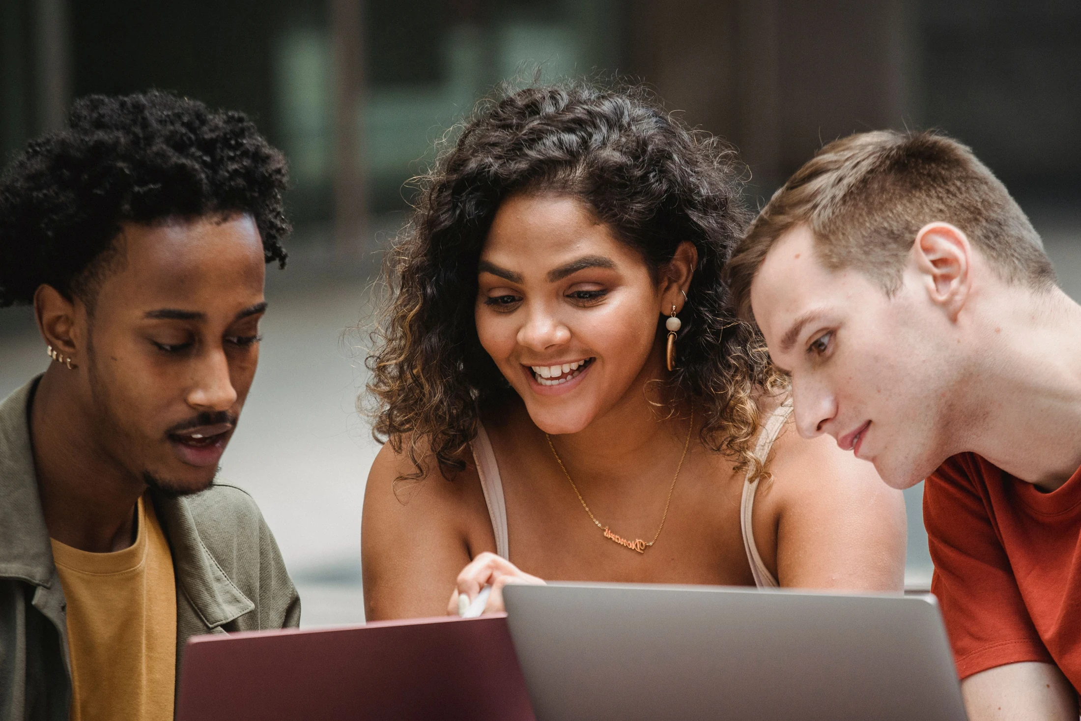 Three young adults lean toward a laptop as they work together outdoors.